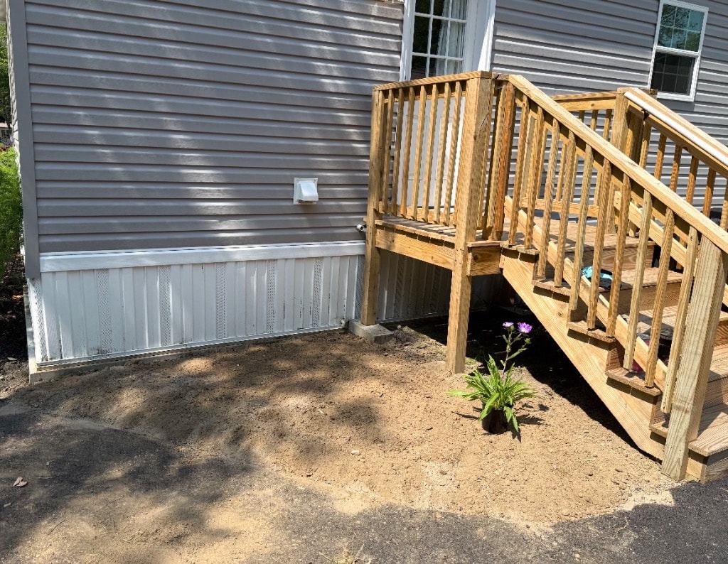 Bare sandy dirt beside front deck stairs with one lone purple flower — Little Garden ready for a shovel