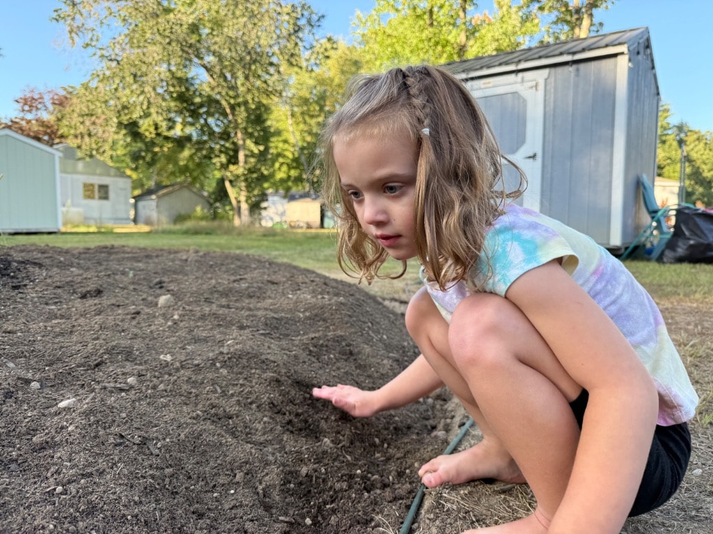 Young girl with braids crouching barefoot over a fresh mound of dark garden soil in the backyard